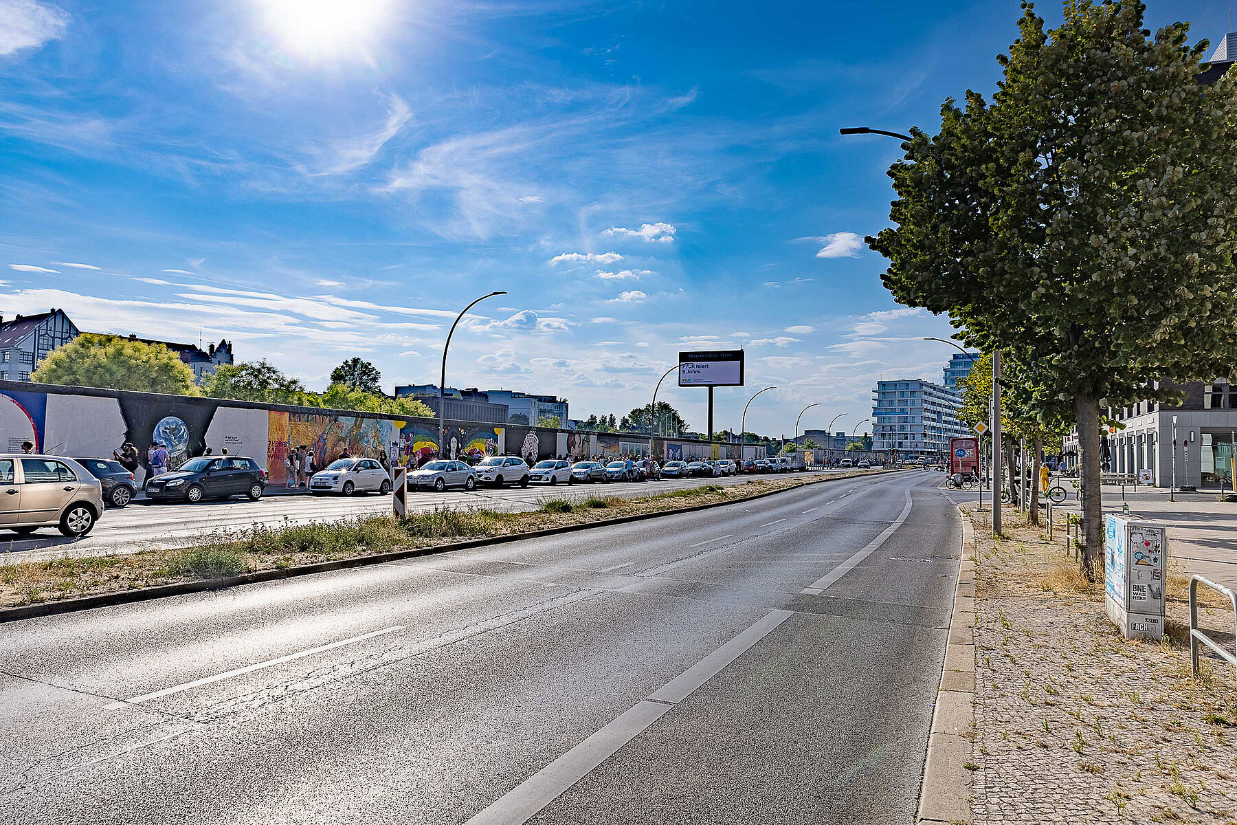 Street with East Side Gallery, cars parked in front of it. There are many street lamps along the street. 