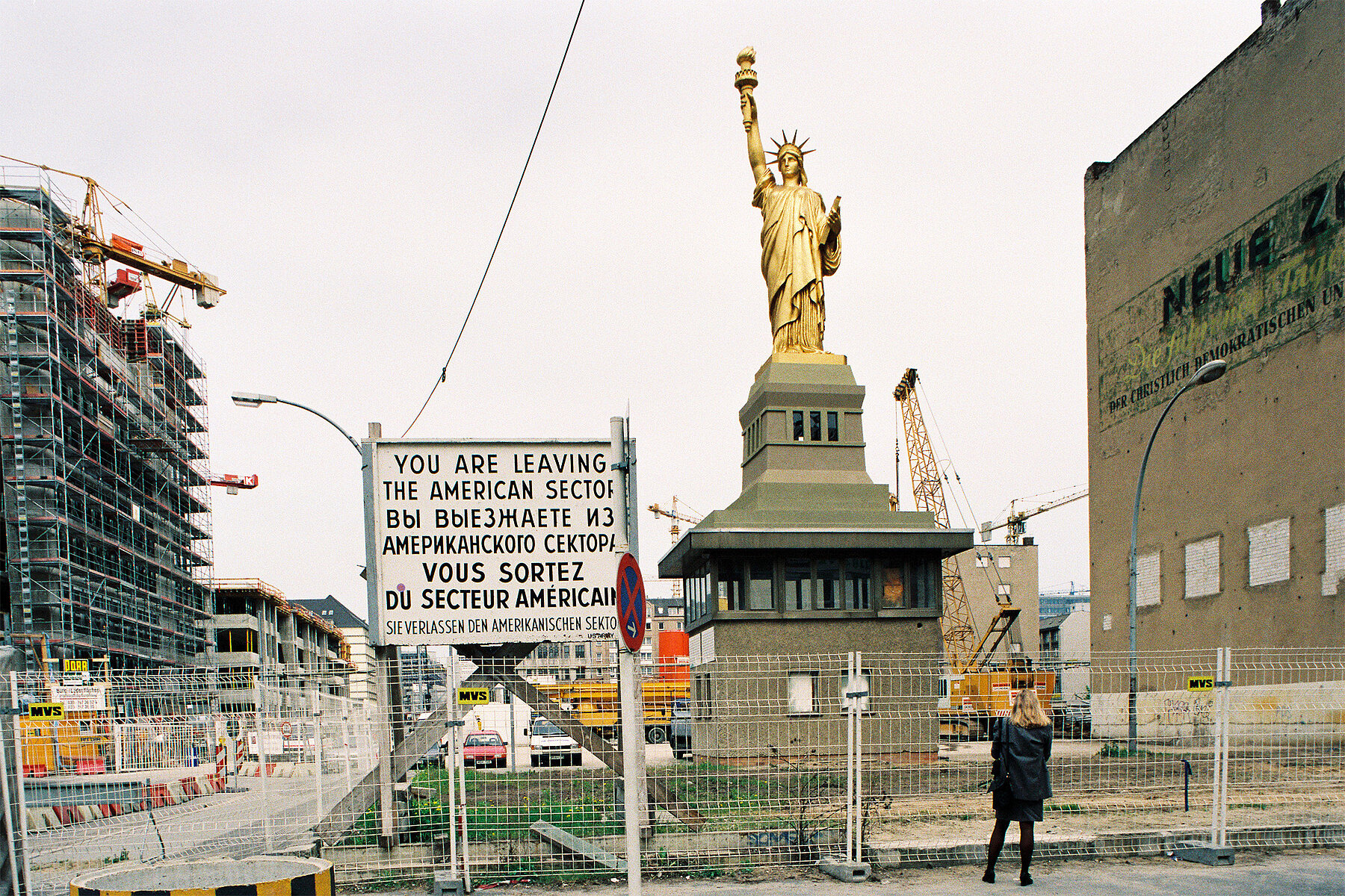 Eine goldene Freiheitsstatue steht auf einem Gebäude. Links ein Schild mit der Aufschrift: Sie verlassen den Amerikanischen Sektor, auf Englisch, Russisch, Französisch und Deutsch. Links hinten befindet sich ein eingerüsteter Neubau.