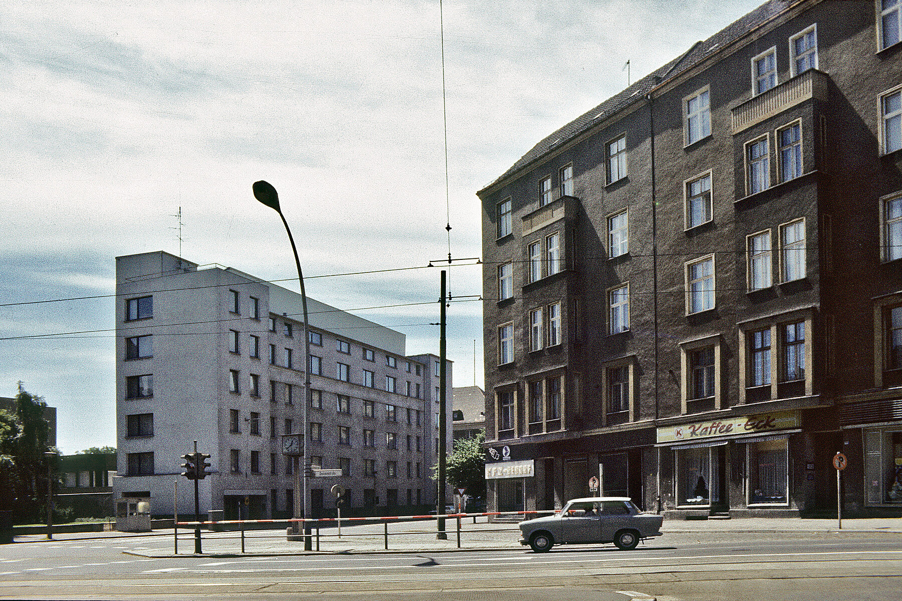 Links das Gebäude der Ständigen Vertretung der Bundesrepublik Deutschland bei der DDR. Rechts ein Gebäude mit Kaffee-Eck und einem vorbeifahrenden Trabi im Vordergrund. 
