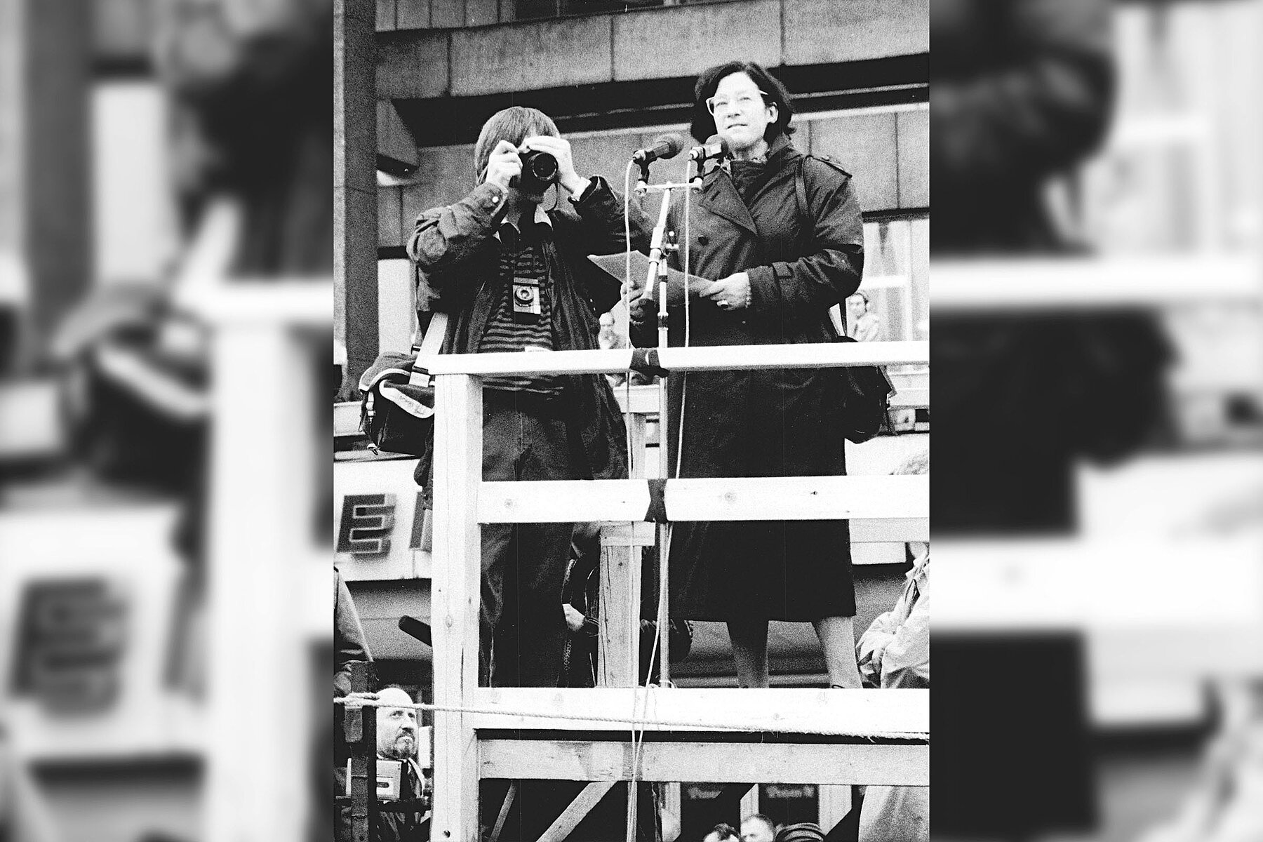 Author Christa Wolf speaks into a microphone on a wooden platform in front of the House of the Teacher. Next to her a man takes photos of the crowd.