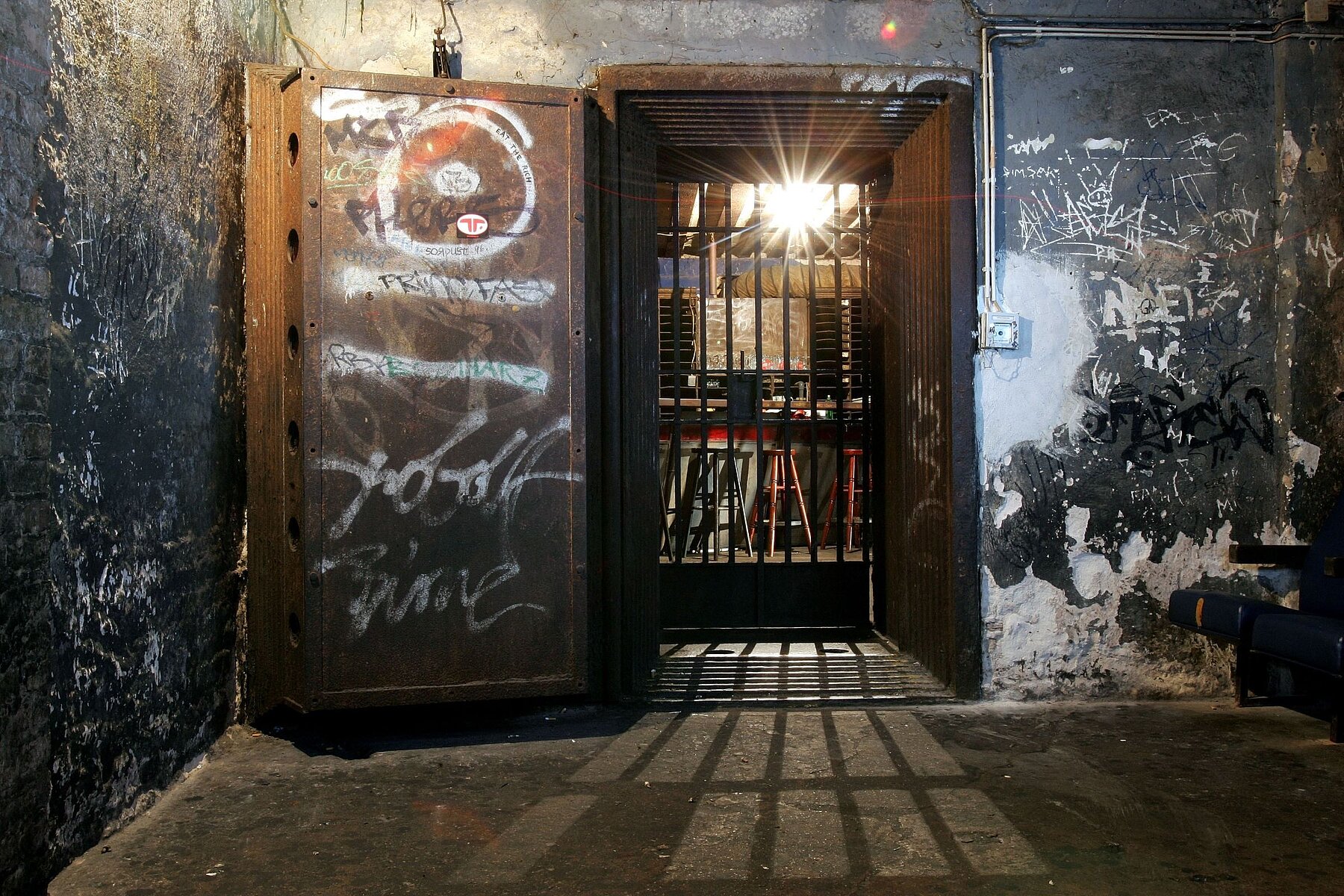 A heavy iron safe door stands open. It is located in a room where the paint is peeling off the walls. Behind the door is a grille through which light shines. 