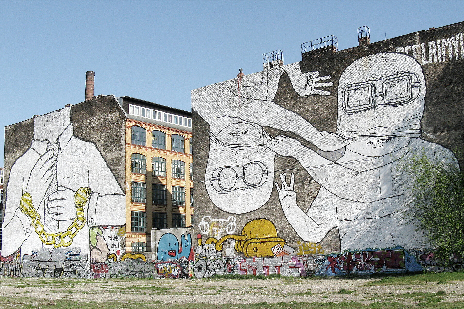 Two graffiti on house walls on the Cuvry wasteland. On the left house there is a headless man wearing gold watches on both wrists, which are connected by a chain. On the right wall are two masked figures wearing diving goggles, the left figure is upside down.