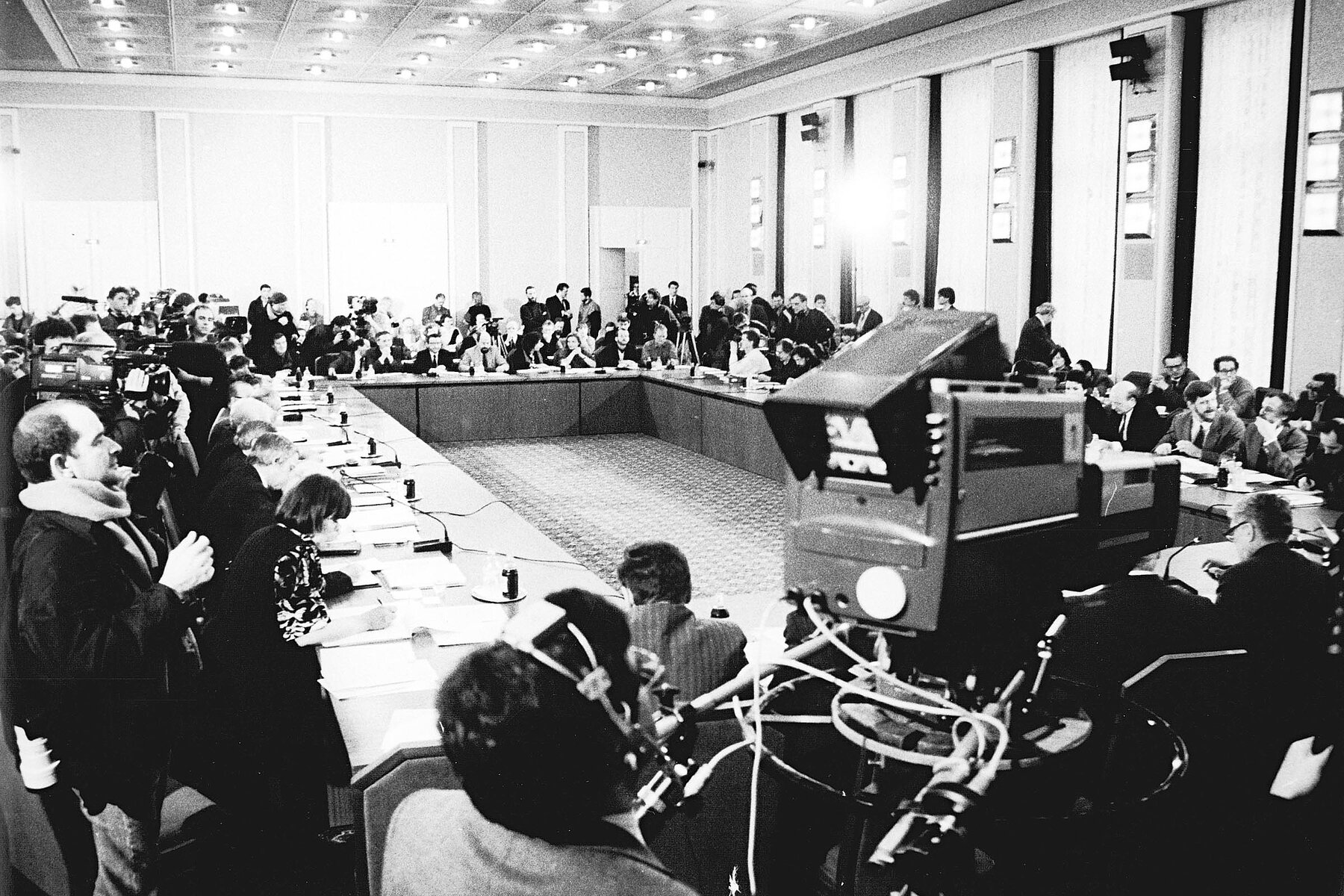 Teilnehmende des Runden Tisches sitzen an im Viereck aufgestellten Tischen im Konferenzsaal vom Schloss Schönhausen, im Vordergrund ragt eine Kamera in das Bild.