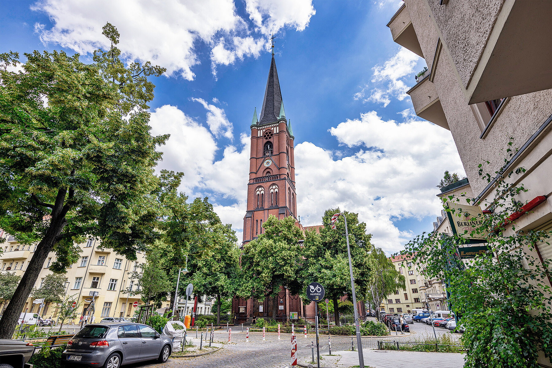 Die Samariterkirche aus rotem Backstein seht auf einer Verkehrsinsel. Sie wird umrahmt von Häusern und Bäumen.