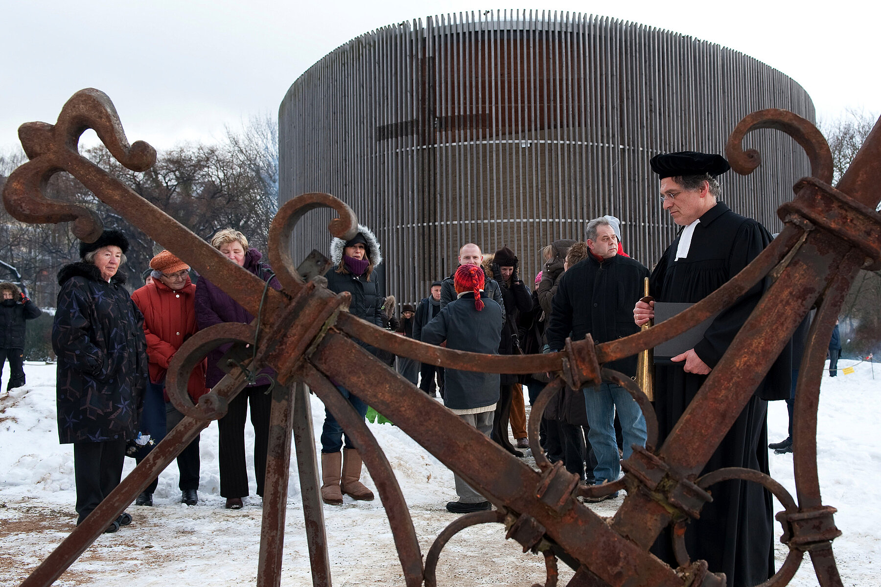 The Chapel of Reconciliation on the grounds of the Berlin Wall Memorial in winter. In front of it, Pastor Manfred Fischer stands with a few other people in the foreground the rusty original cross of the blown-up Church of Reconciliation.